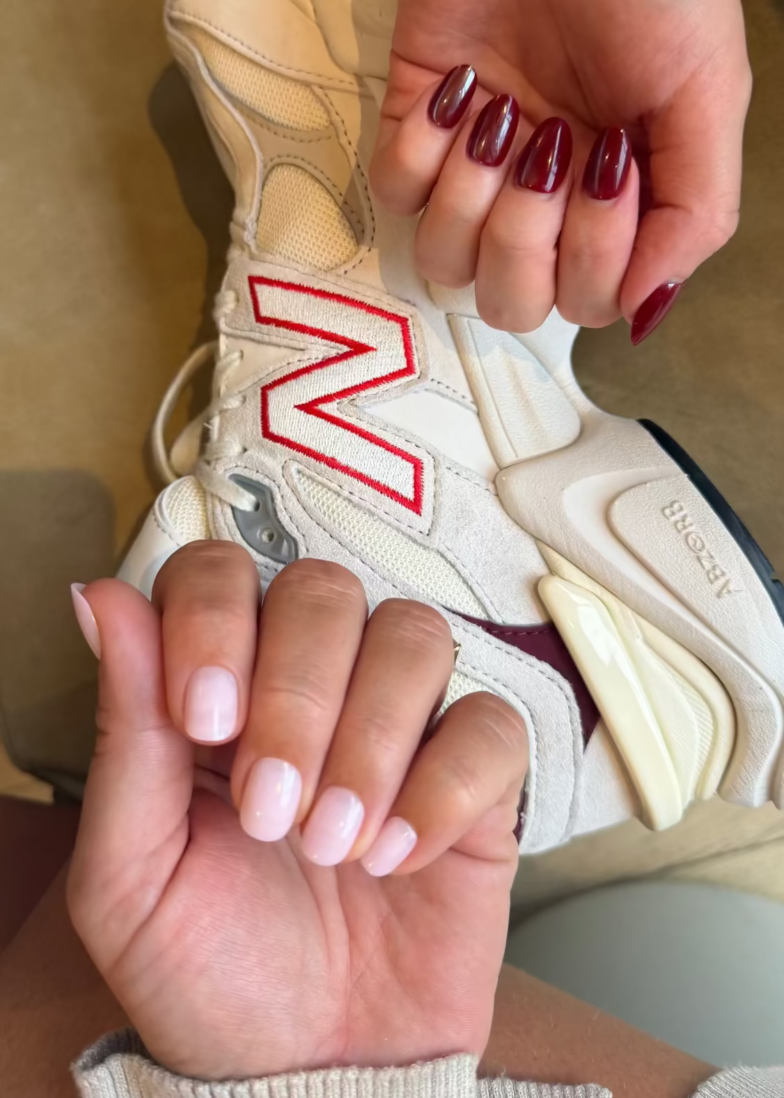 Person holding a beige sneaker with a red logo, wearing maroon nail polish.